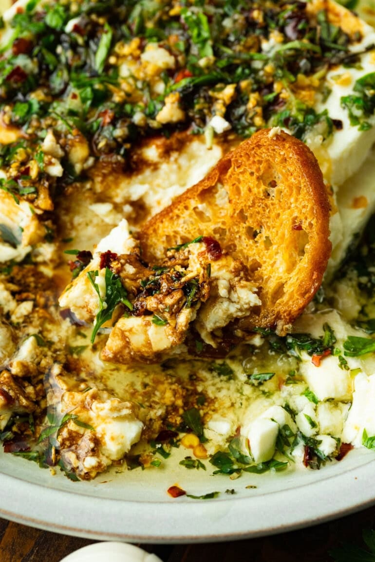a photo of a crostini dipped in a bowl of creamy feta topped with hot herb oil