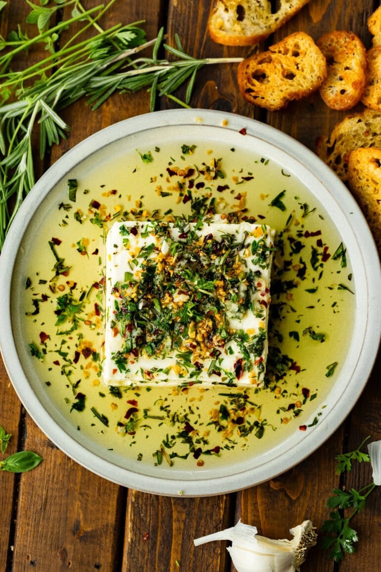 a square brick of feta cheese sitting on a serving plate in a pool of herby olive oil
