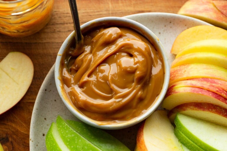 A bowl of caramel has a big serving spoon in it and is surrounded by sliced apples