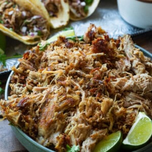 a photo of a bowl of shredded pork carnitas with three tacos in the background