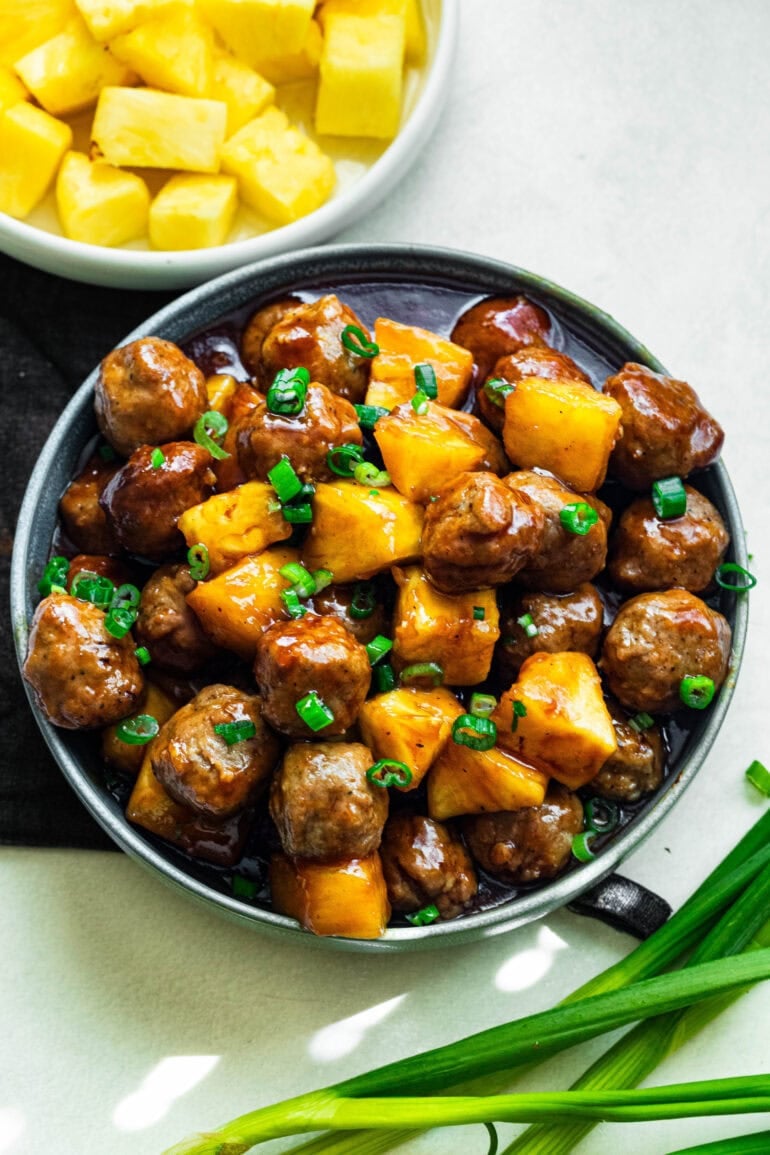 a large serving bowl of saucy hawaiian meatballs mixed with pineapple chunks and topped with chopped green onions