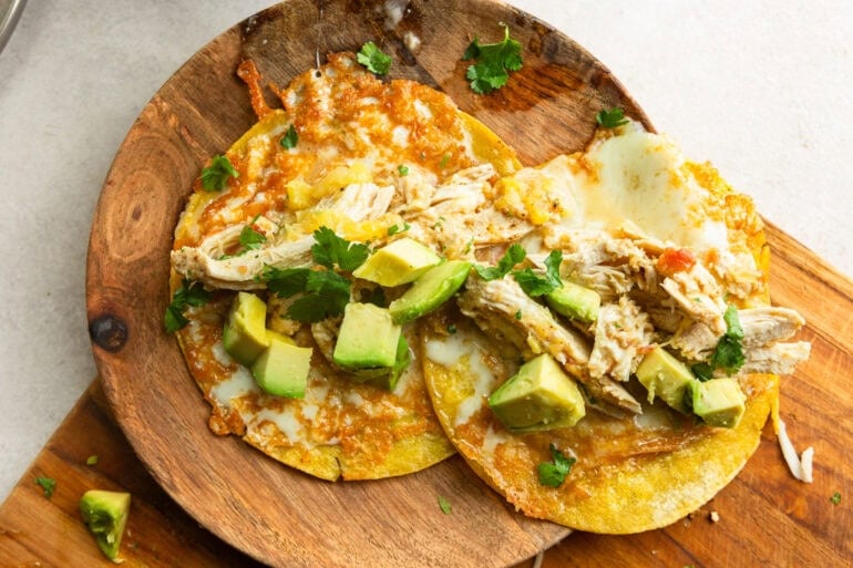 A wooden cutting board has two shredded chicken tacos on yellow corn tortillas with melty cheese, shredded chicken, avocado, and cilantro.