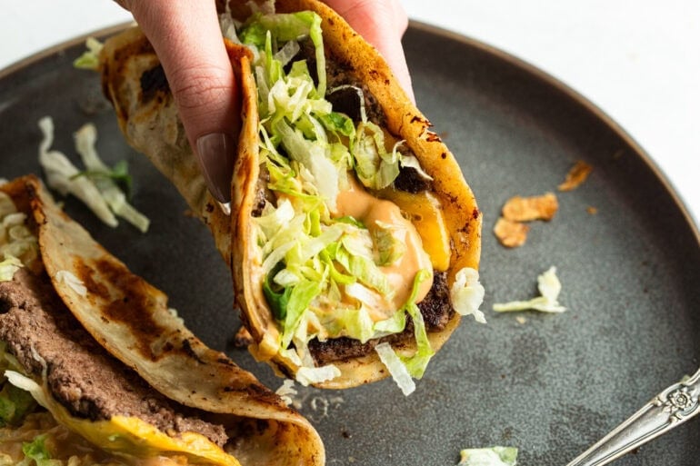 someone holding a smash burger taco over a plate loaded with crispy beef, shredded lettuce, melted cheese and a burger sauce