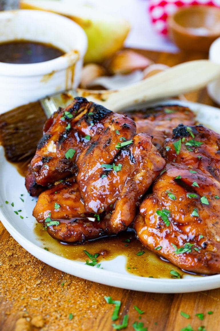 A plate with marinated, smoked chicken thighs. A brush dripping with marinade is next to the chicken and a bowl of marinade is in the background.
