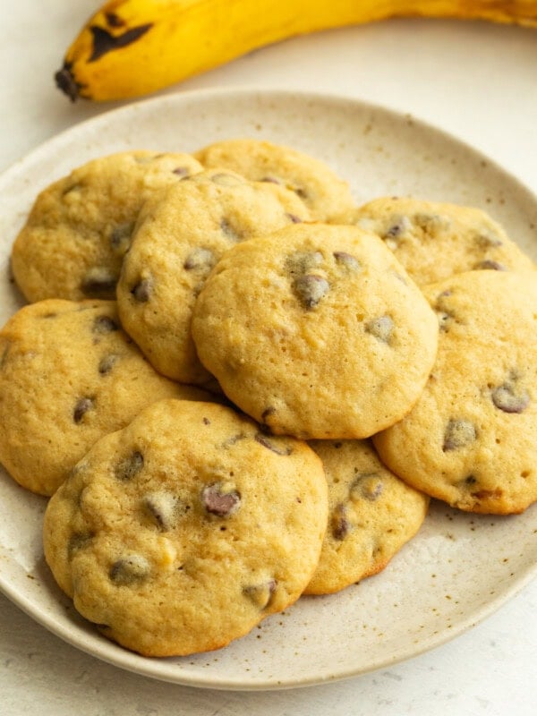 Soft chocolate chip cookies on white ceramic plate with banana in background.
