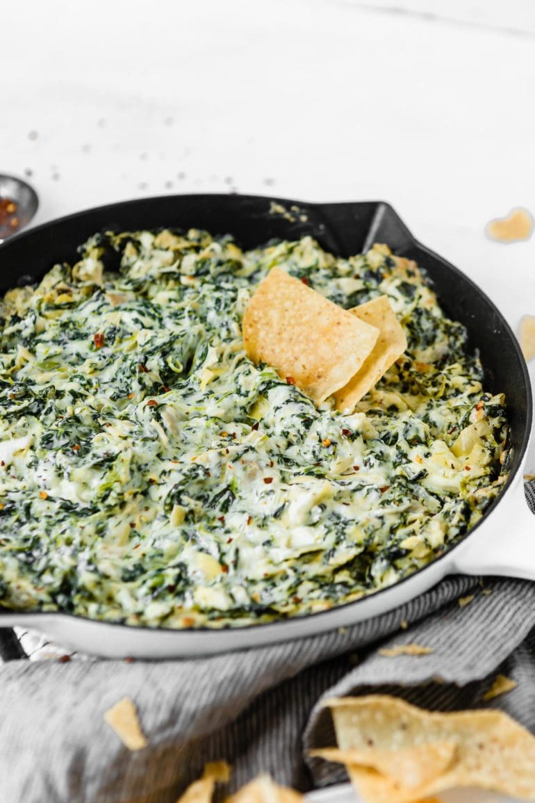A cast iron skillet filled with spinach and artichoke dip. You can see the creamy white cheese sauce with green spinach and artichokes. There are two tortilla chips in the dip.