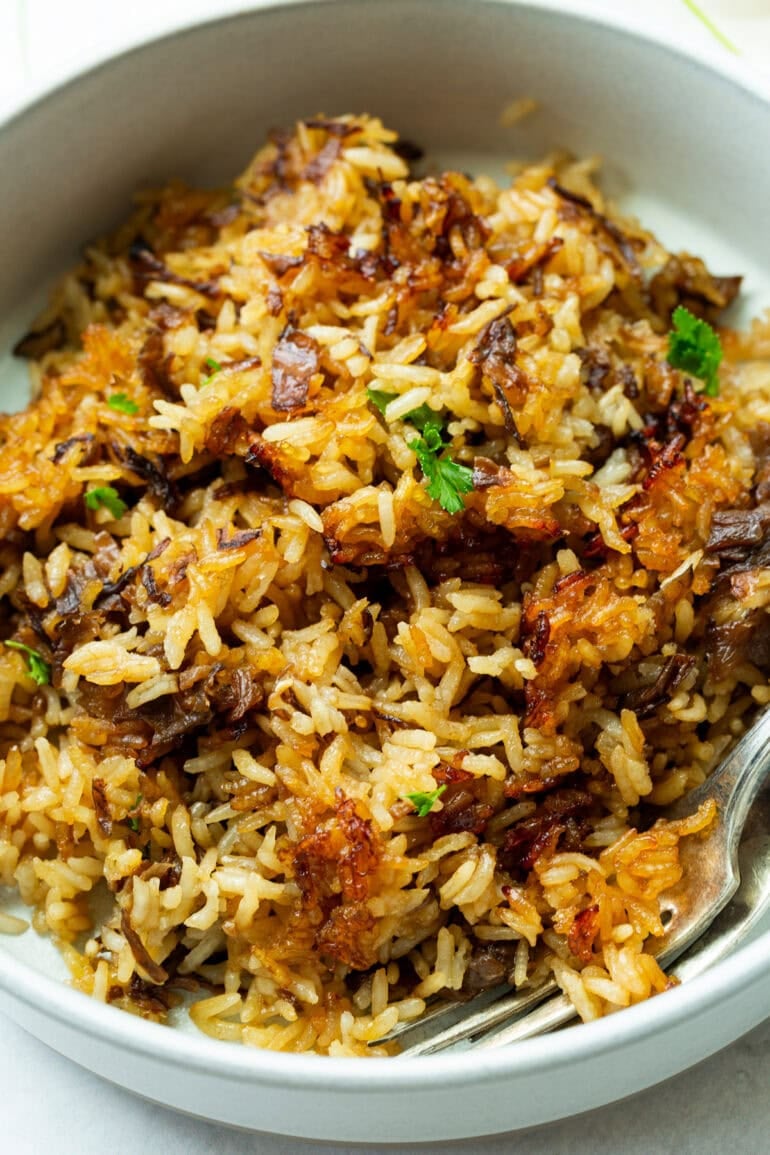 A close-up shot of Stick of Butter Rice with Beef Broth in a white bowl. The grains are fluffy and tender, with visible bits of caramelized onion from the soup mix. The rich brown color hints at the beef broth and buttery flavor.