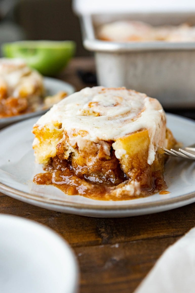 a photo of a single sticky apple cinnamon roll sitting on a white saucer topped with browned butter icing and sitting in a pool of caramel sticky sauce.