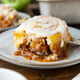 a photo of a single sticky apple cinnamon roll sitting on a white saucer topped with browned butter icing and sitting in a pool of caramel sticky sauce.