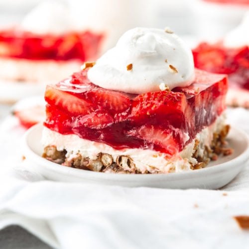 A serving of strawberry pretzel jello salad on a white dessert plate. The salad is layered with pretzel on the bottom, then a white cream layer topped with a layer of strawberry jello packed with sliced strawberries. A dollop of cool whip is on top. Two other servings are in the background.