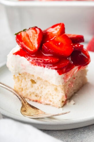 a photo of a single piece of moist white cake on a small dessert plate topped with whipped cream cream cheese frosting and juicy red strawberries