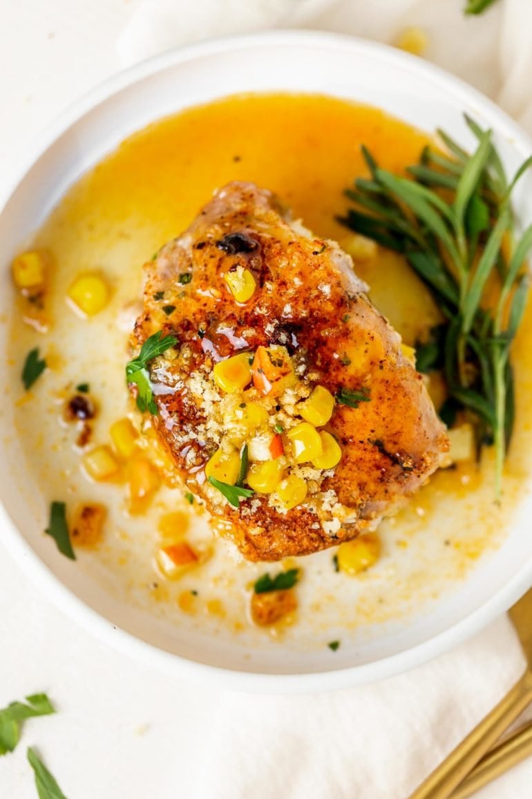 A stuffed pork chop on a dinner plate. There is a little stuffing with corn kernels on top of the pork chop and a sauce has been poured over the top. Fresh sage is on the edge of the plate.