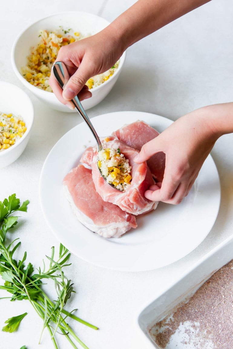 Uncooked pork chops. One pork chop has been cut opened and a spoonful of stuffing is being put inside. Fresh sage and a bowl of bread crumbs are sitting nearby.