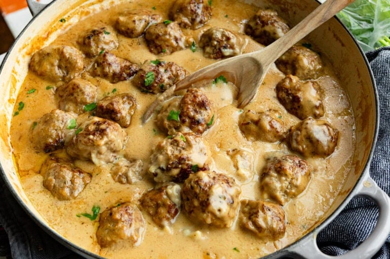 swedish meatballs sitting in thick gravy sauce being stirred with a wooden spoon
