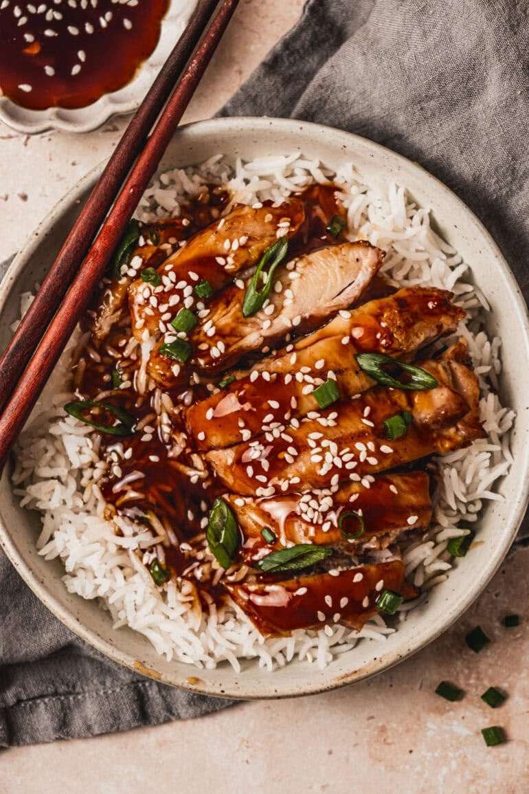 a plate of juicy teriyaki chicken thighs sliced on top of rice and sprinkled with sesame seeds and green onions with chopsticks sitting on the side of the plate