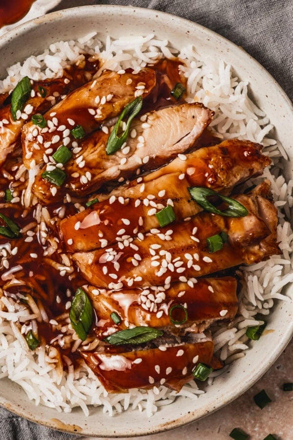 several slices of juicy chicken thigh topped with teriyaki sauce over a bed of white rice and sprinkled with sesame seeds and sliced green onions