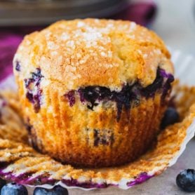 A blueberry muffin with the paper liner peeled back so you can see the golden brown muffin packed with blueberries.