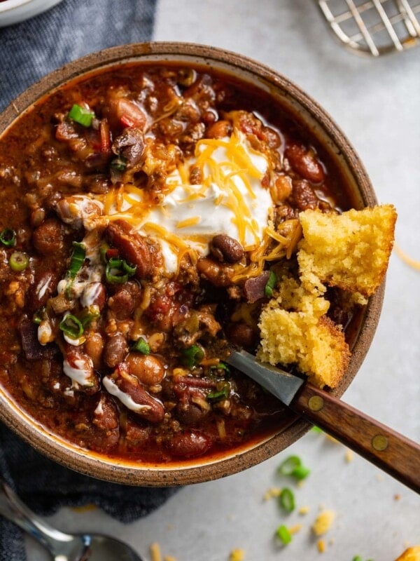 A bowl of chili. The chili has ground beef, beans and a rich sauce. It is topped with sour cream and shredded cheese and a piece of cornbread is on the edge of the bowl.