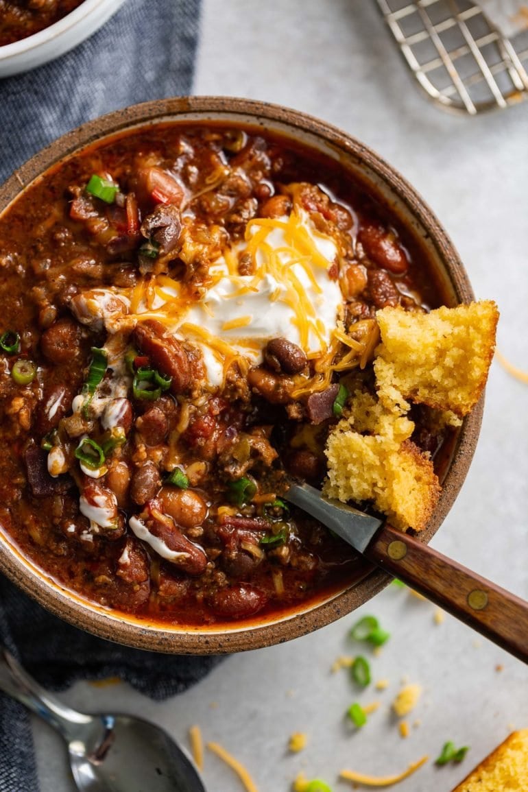 A bowl of chili. The chili has ground beef, beans and a rich sauce. It is topped with sour cream and shredded cheese and a piece of cornbread is on the edge of the bowl.