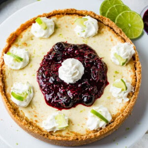 An aerial photo of a whole cheesecake on a white serving platter with dollops of whipped cream around the edges with a slice of lime in each dollop and a pile of triple berry sauce in the middle with a dollop of whipped cream on top