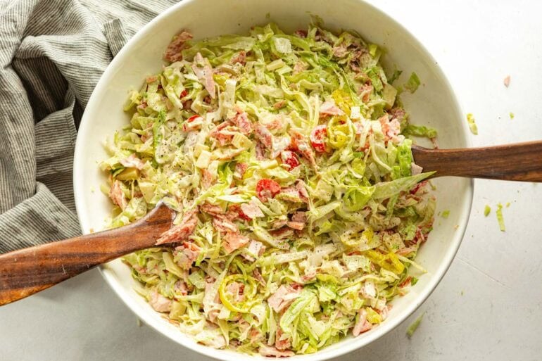 a grinder salad being tossed with wooden spoons in a large white bowl