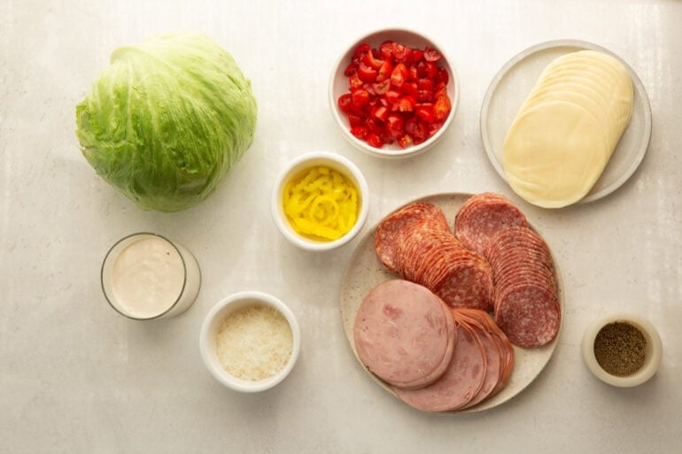 all the ingredients for a grinder salad including salami, ham, provolone, icebery lettuce, red bell pepper, and seasonings