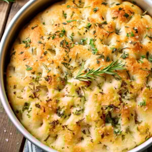 A photo of a round focaccia bread loaf covered in fresh herbs and dusted with salt flakes in a silver pan.