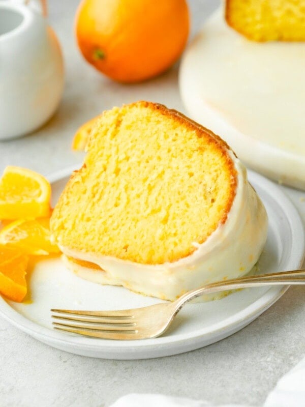 a photo of a slice of bright orange bundt cake topped with a white glaze sitting on a white plate with a fork in front of if and small orange slices scattered around it.