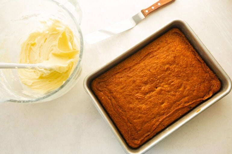 a baked 9x13 carrot cake with a bowl of cream cheese frosting in a glass bowl with an offset spatula sitting next to the bowl