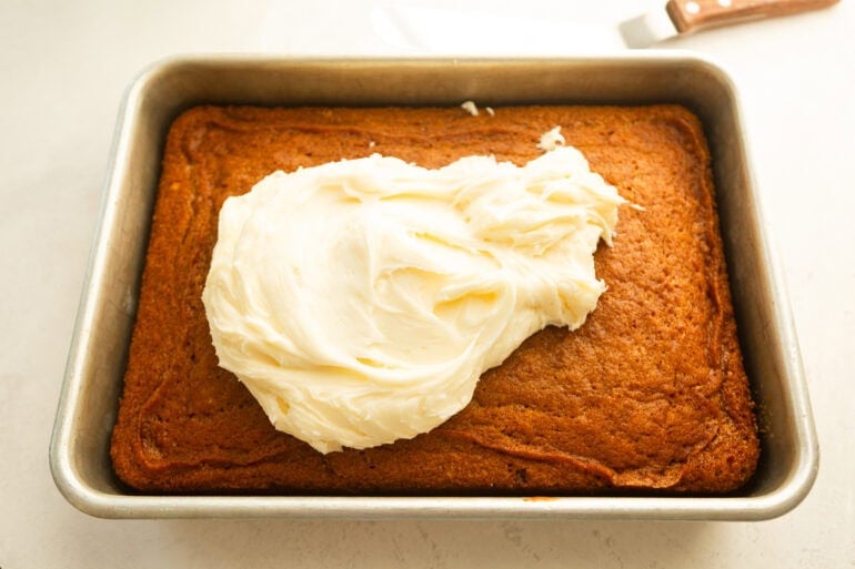 a baked carrot cake in a a 9x13 baking pan with cream cheese frosting piled on top ready to be spread out