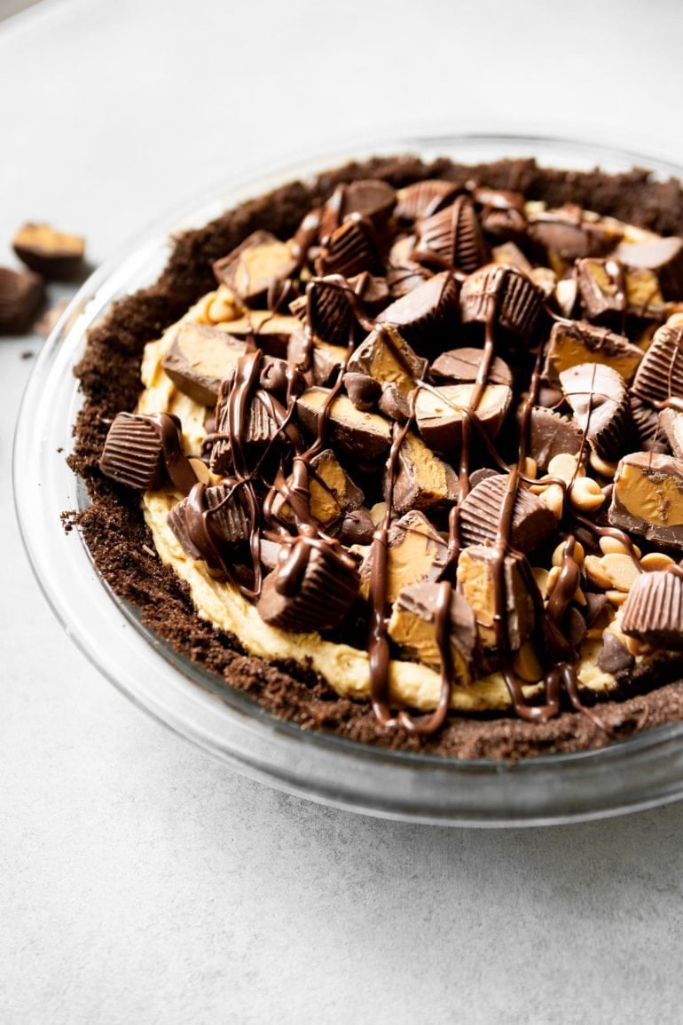 a photo of a peanut butter pie with a chocolate graham cracker crust topped with mini peanut butter cupes, chocolate chips and peanut butter chips with a chocolate drizzle.