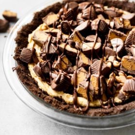a photo of a peanut butter pie with a chocolate graham cracker crust topped with mini peanut butter cupes, chocolate chips and peanut butter chips with a chocolate drizzle.