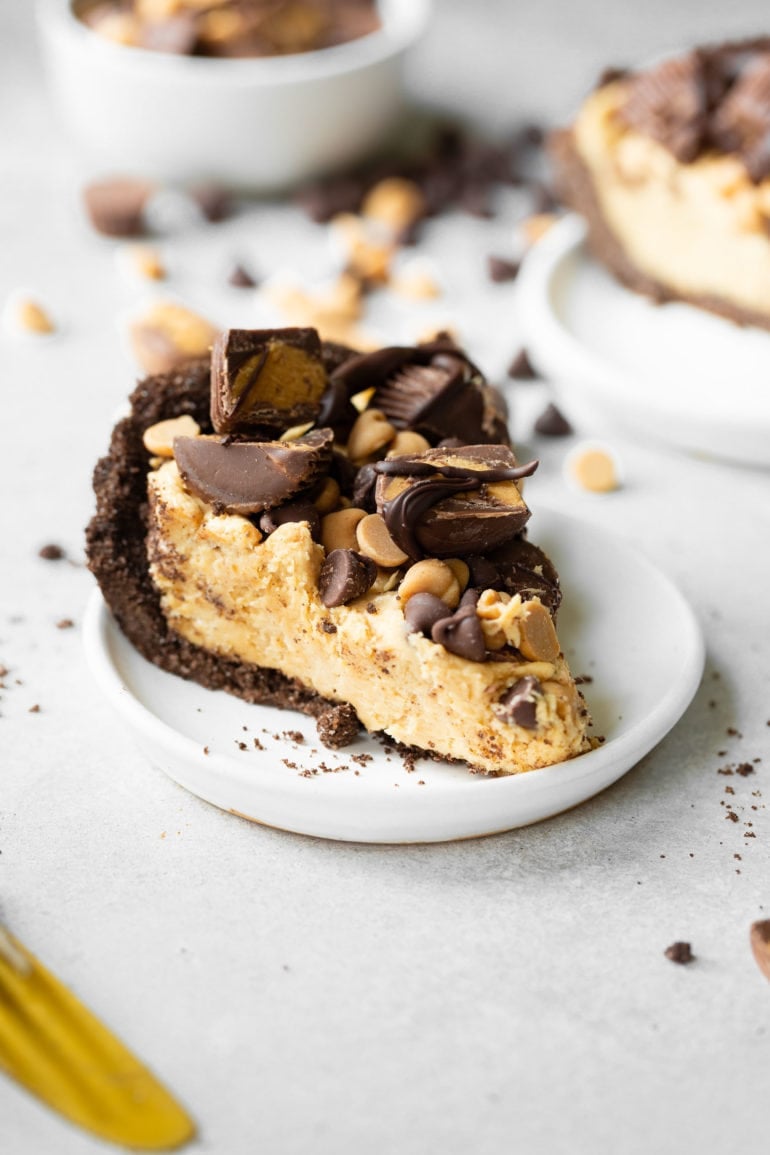 a photo of a slice of peanut butter pie on a small white saucer. the pie is topped with peanut butter cups and chocolate chips and peanut butter chips.