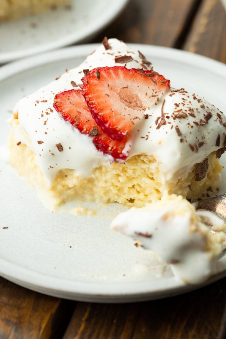 a photo of a moist piece of tres leches cake topped with sliced strawberries and shaved chocolate with a bite removed by a fork