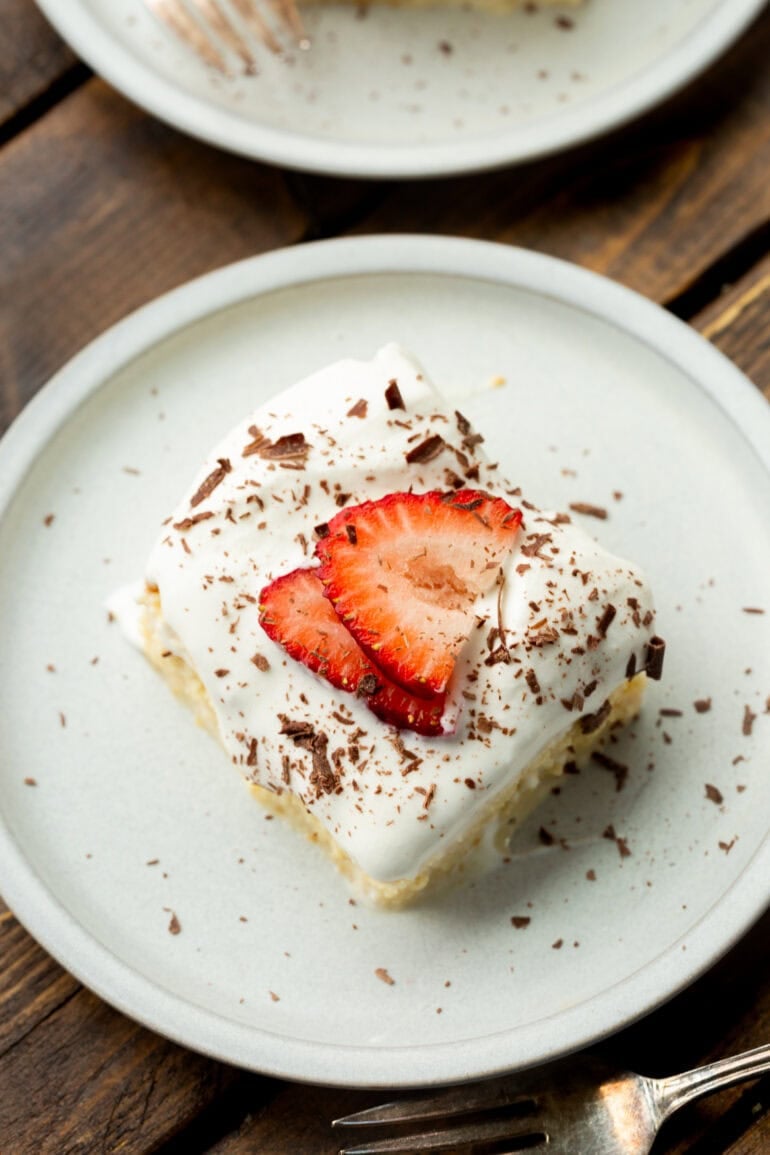 a photo taken over the top of a piece of tres leches cake on a small white dessert plate topped with sliced strawberries and shaved chocolate