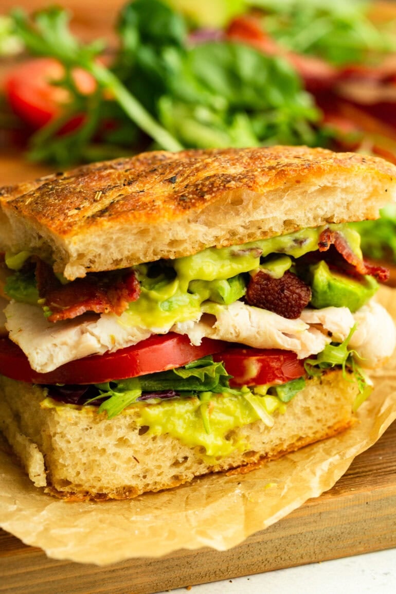 half of a turkey bacon avocado sandwich on focaccia great with avocado sauce, tomatoes, lettuce, thick turkey slices and crispy bacon