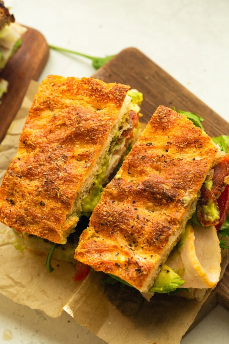 turkey bacon avocado sandwich on focaccia bread cut in half and sitting on a piece of brown parchment paper