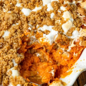 a photo of a round baking dish full of sweet potato casserole topped with gooey marshmallows and a brown sugar streusel topping with a serving being scooped out of the dish