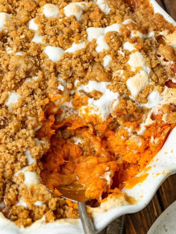 a photo of a round baking dish full of sweet potato casserole topped with gooey marshmallows and a brown sugar streusel topping with a serving being scooped out of the dish