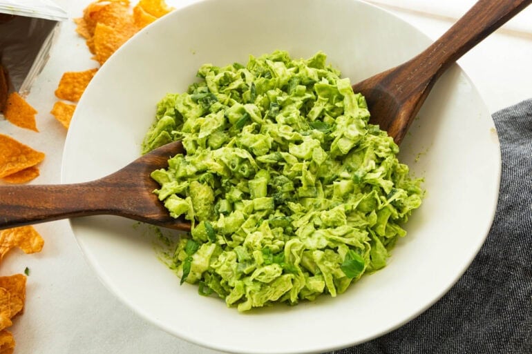 a large white serving bowl filled with green goddess salad with two large wooden spoons about to toss it