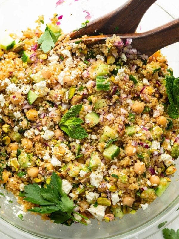 a large glass bowl full of a chickpea, quinoa and cucumber salad with red onions and feta cheese