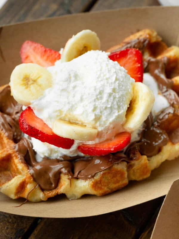 a photo of a golden liege waffle in a brown cardboard carton topped with nutella, sliced strawberries and bananas and whipped cream
