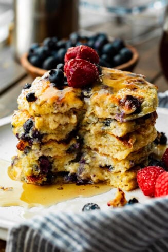 a photo of a stack 5 fluffy blueberry pancakes with a bite taken out topped with fresh berries and covered with syrup