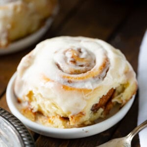 a photo of a single cinnamon roll topped with a creamy white frosting sitting on a small dessert plate with a silver fork in the foreground.