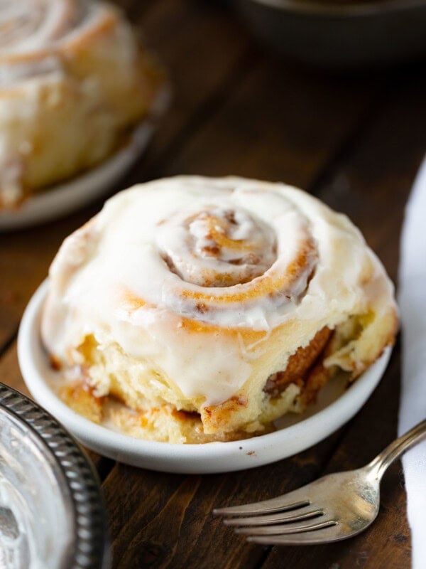 a photo of a single cinnamon roll topped with a creamy white frosting sitting on a small dessert plate with a silver fork in the foreground.