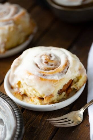 a photo of a single cinnamon roll topped with a creamy white frosting sitting on a small dessert plate with a silver fork in the foreground.
